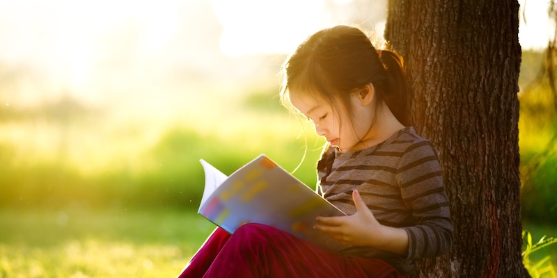 young girl reading a book against a tree